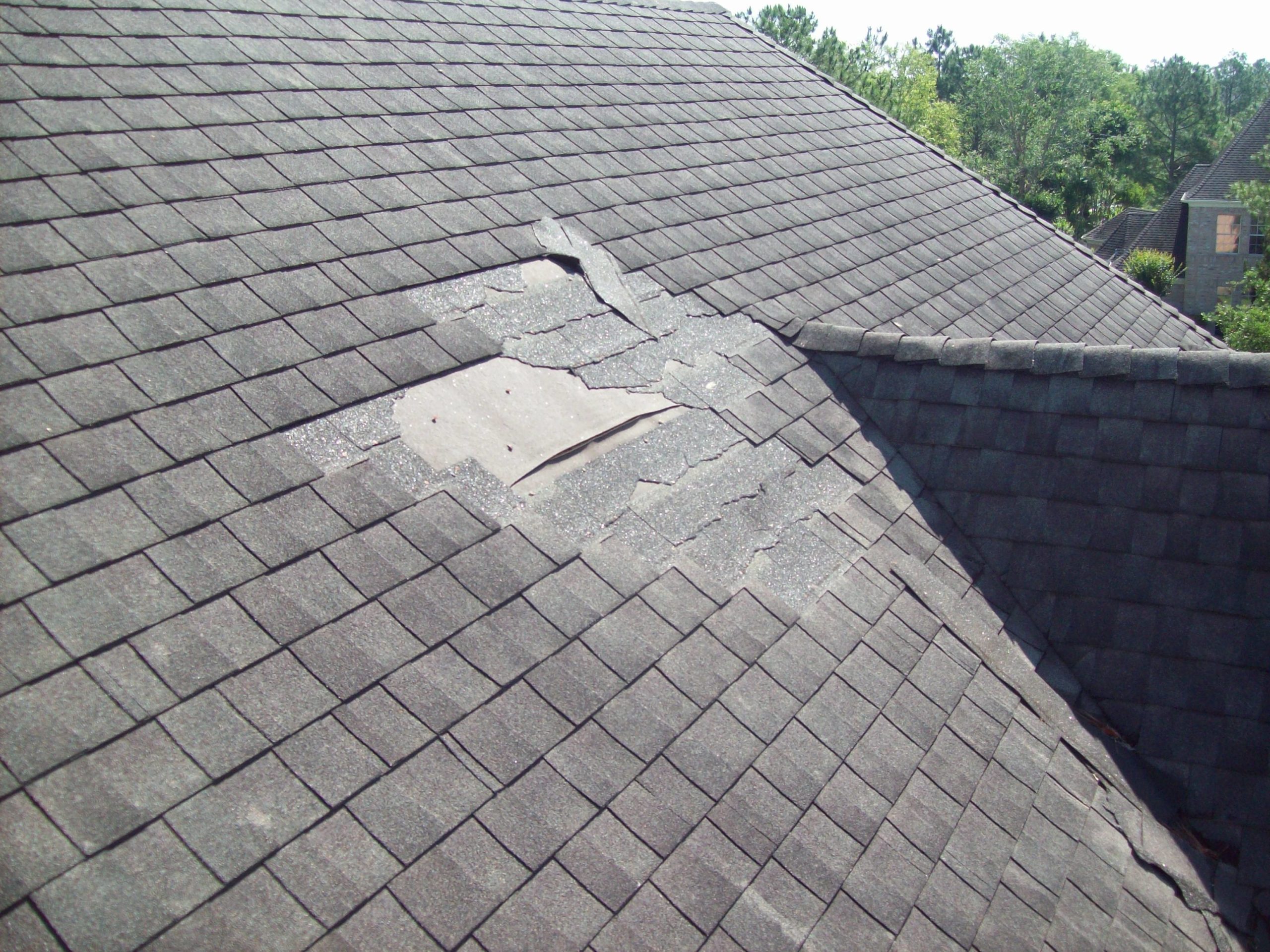 An image of a residential roof with gray asohalt shingles. The shingles have been damaged in a storm and some are missing.