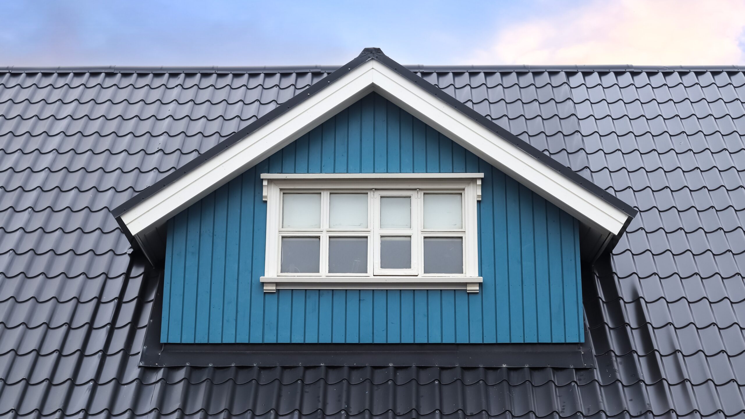Home with blue siding on roof gable with dark steel roofing and a beautiful sky view behind it