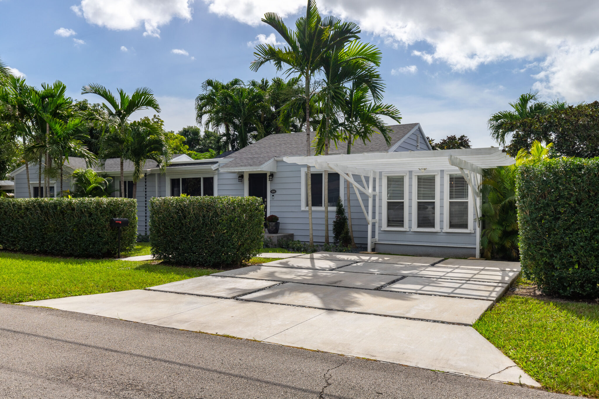 Pale blue home with black asphalt roof surrounded by greenery and palm trees