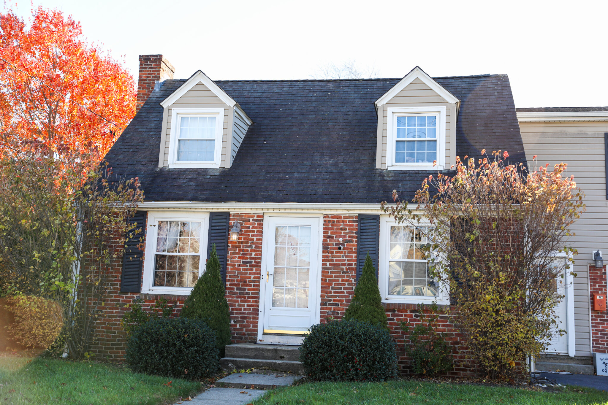 The image features a smaller brick house with black asphalt shingles. There are trees and shrubbery surrounding the home.