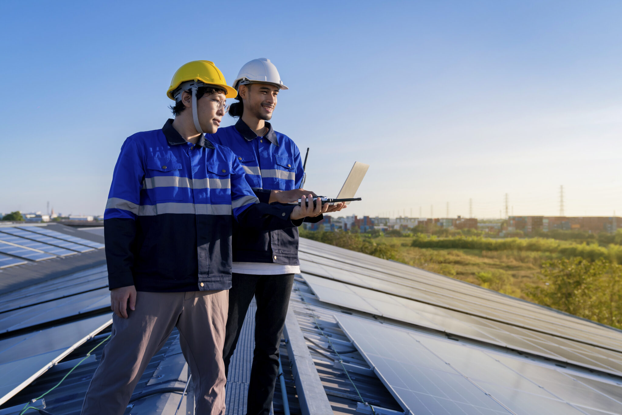 Two smiling young men who are roofing technicians standing on top of the solar panel and metal tile roof that they are installing with an open laptop