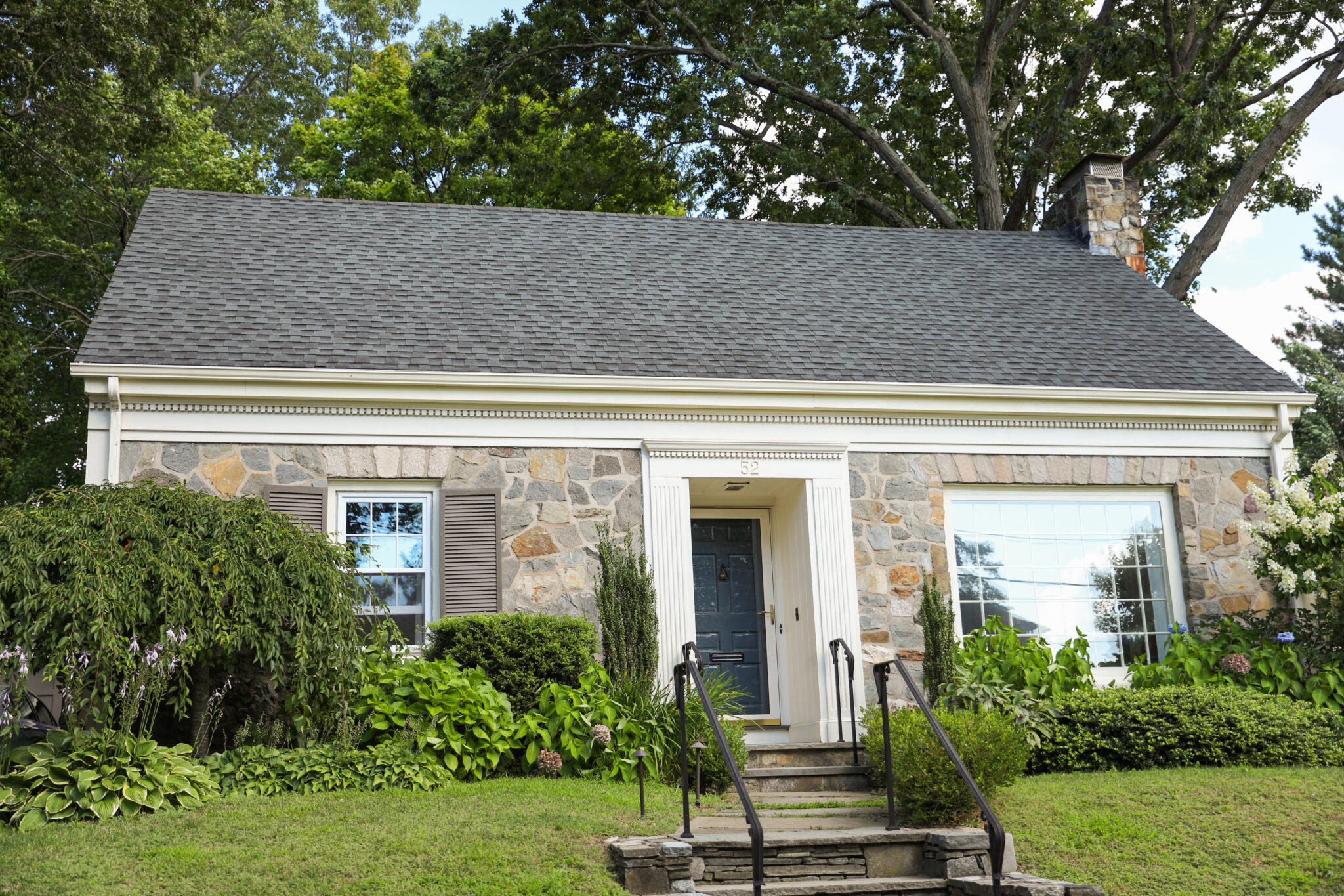 This image features a beautiful brick house with new asphalt shingles. The yard is small, but very cute with vibrant grass, trees, and bushes.