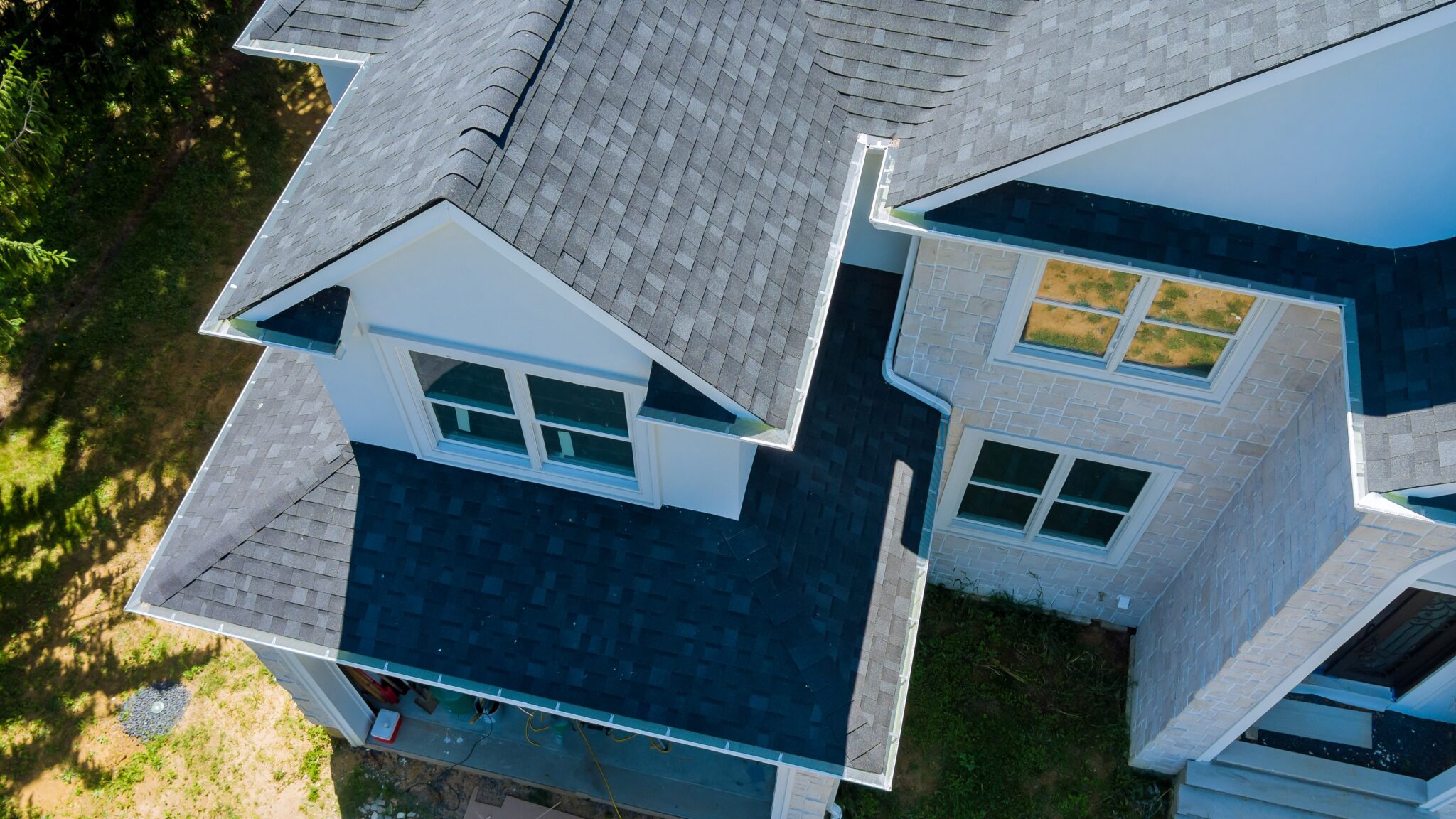 A drone shot of a home exterior with a dark asphalt shingle roof, faux brick siding, windows with white frames, and white gutters